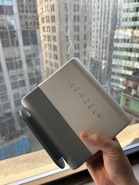 Hand holding a white device with buttons, against a city window backdrop.