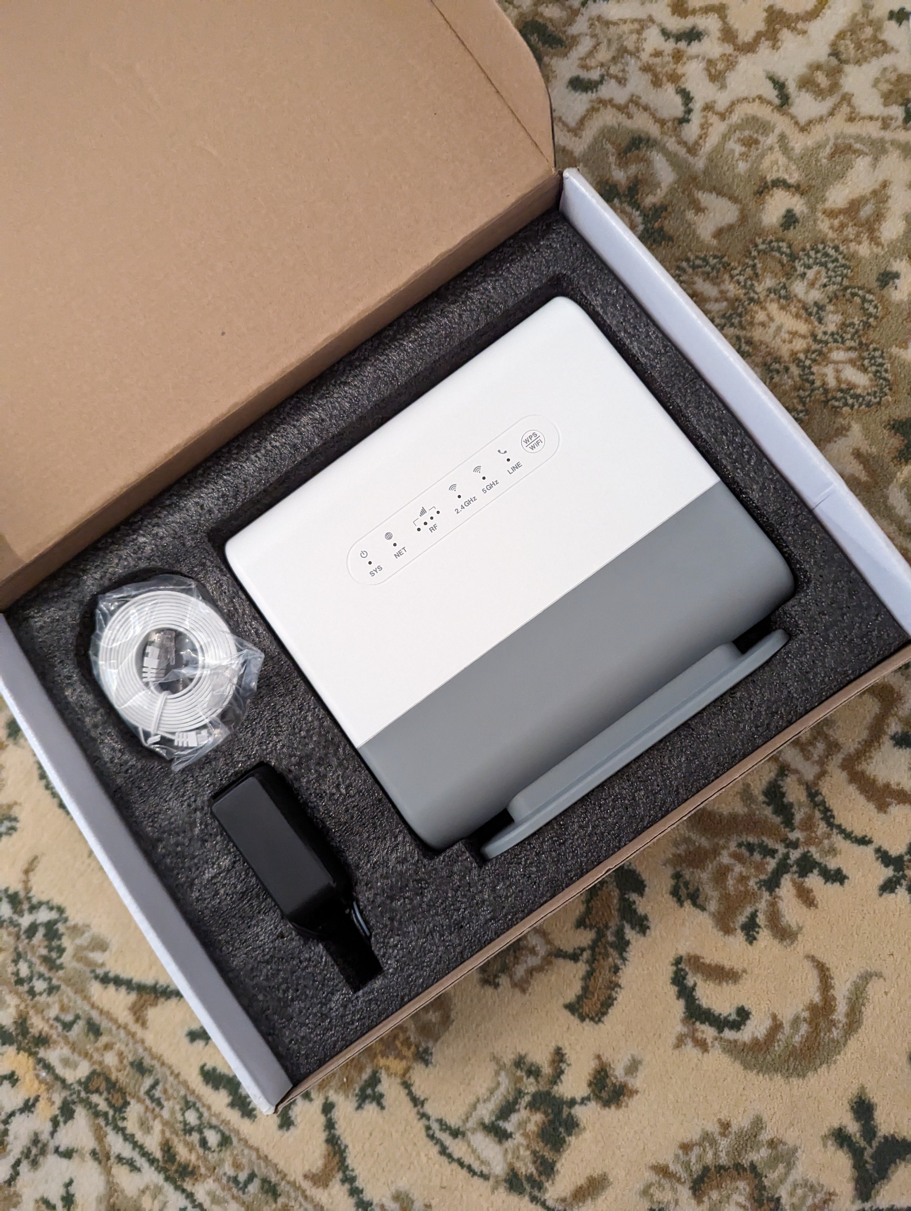 Router and accessories packaged in a cardboard box on a patterned carpet.