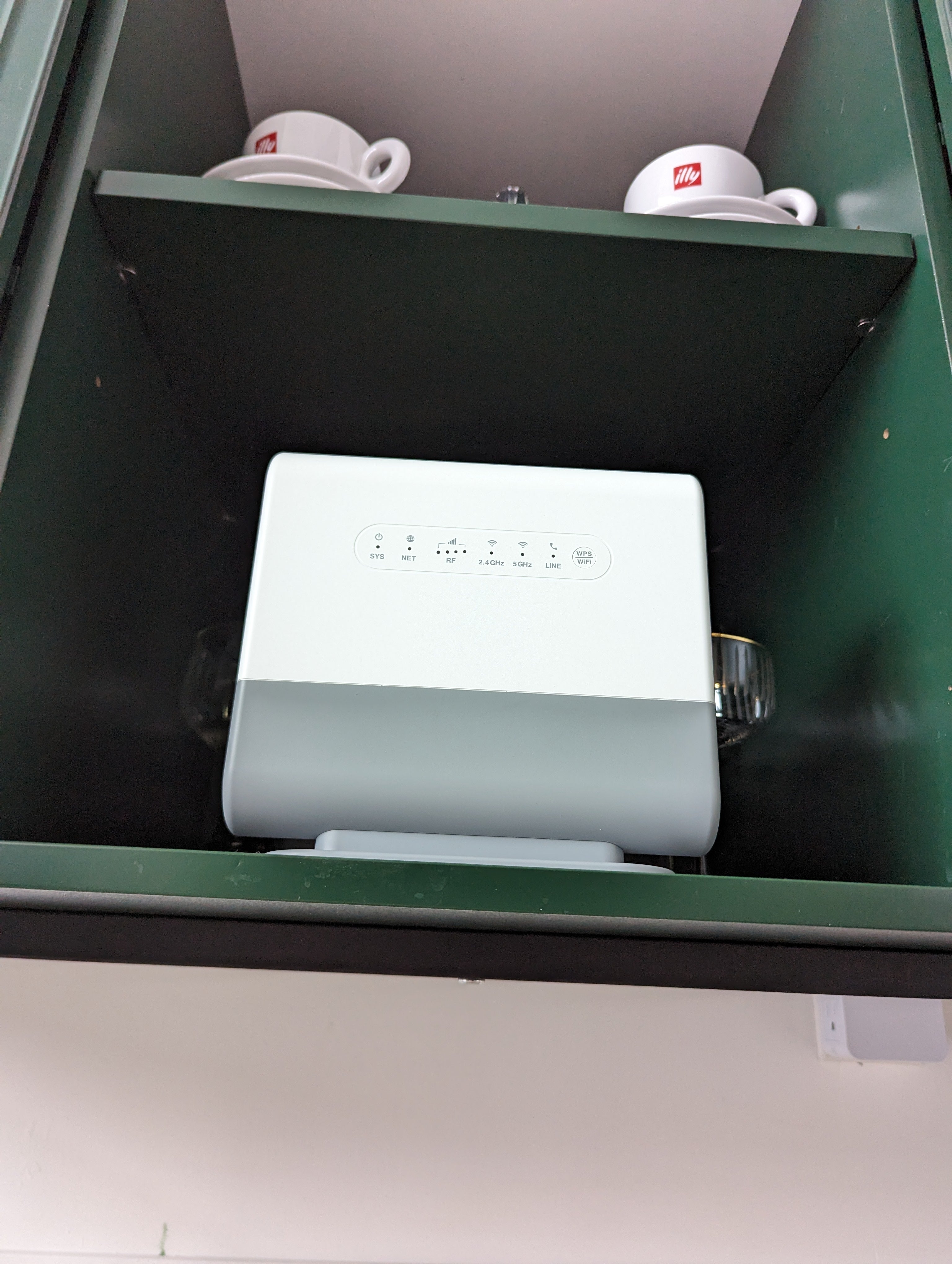 White router inside a green cupboard, with two coffee cups on the shelf above.