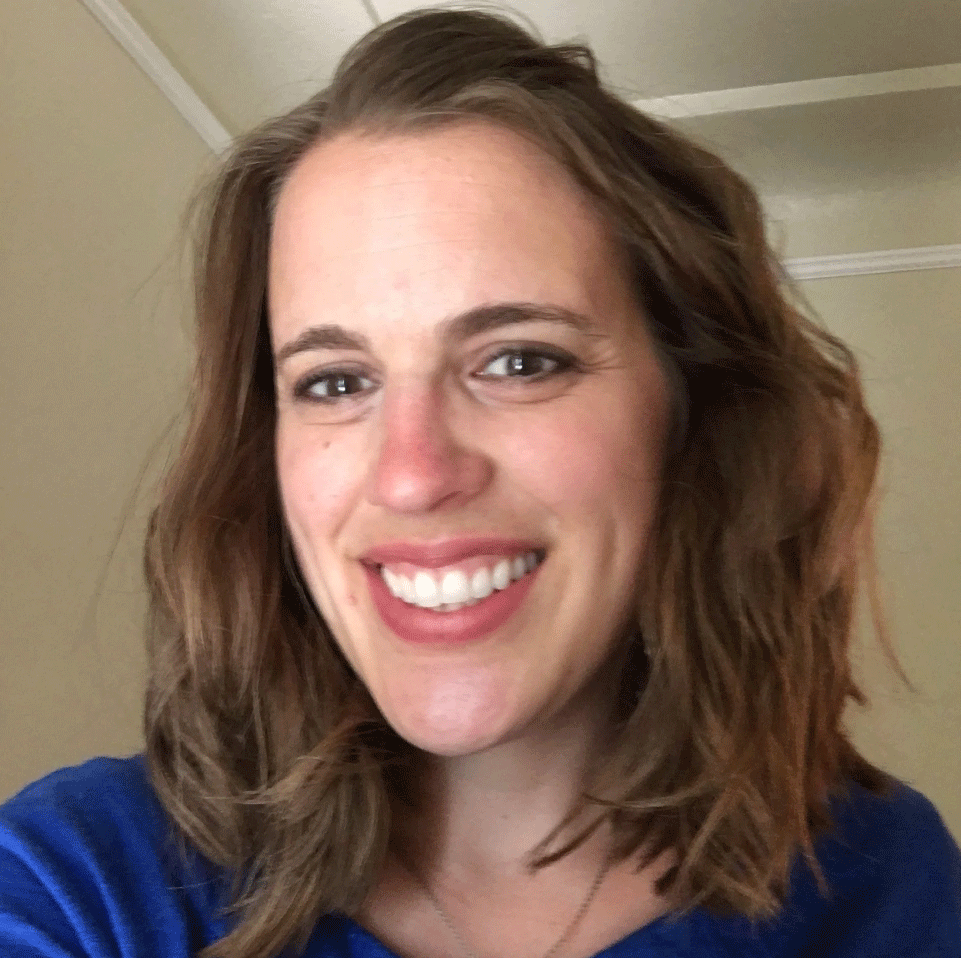 A woman in a purple shirt smiling, seated outdoors by a wooden fence.