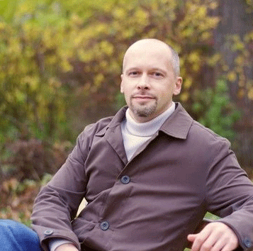 Man sitting outdoors in a brown coat, surrounded by fall foliage.